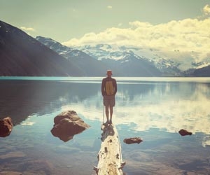 Man standing on log in lake with mountains reflecting on water